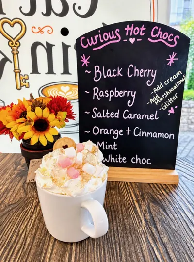 A hot chocolate piled with cream and marshmallows beside our Curious Hot Chocs board